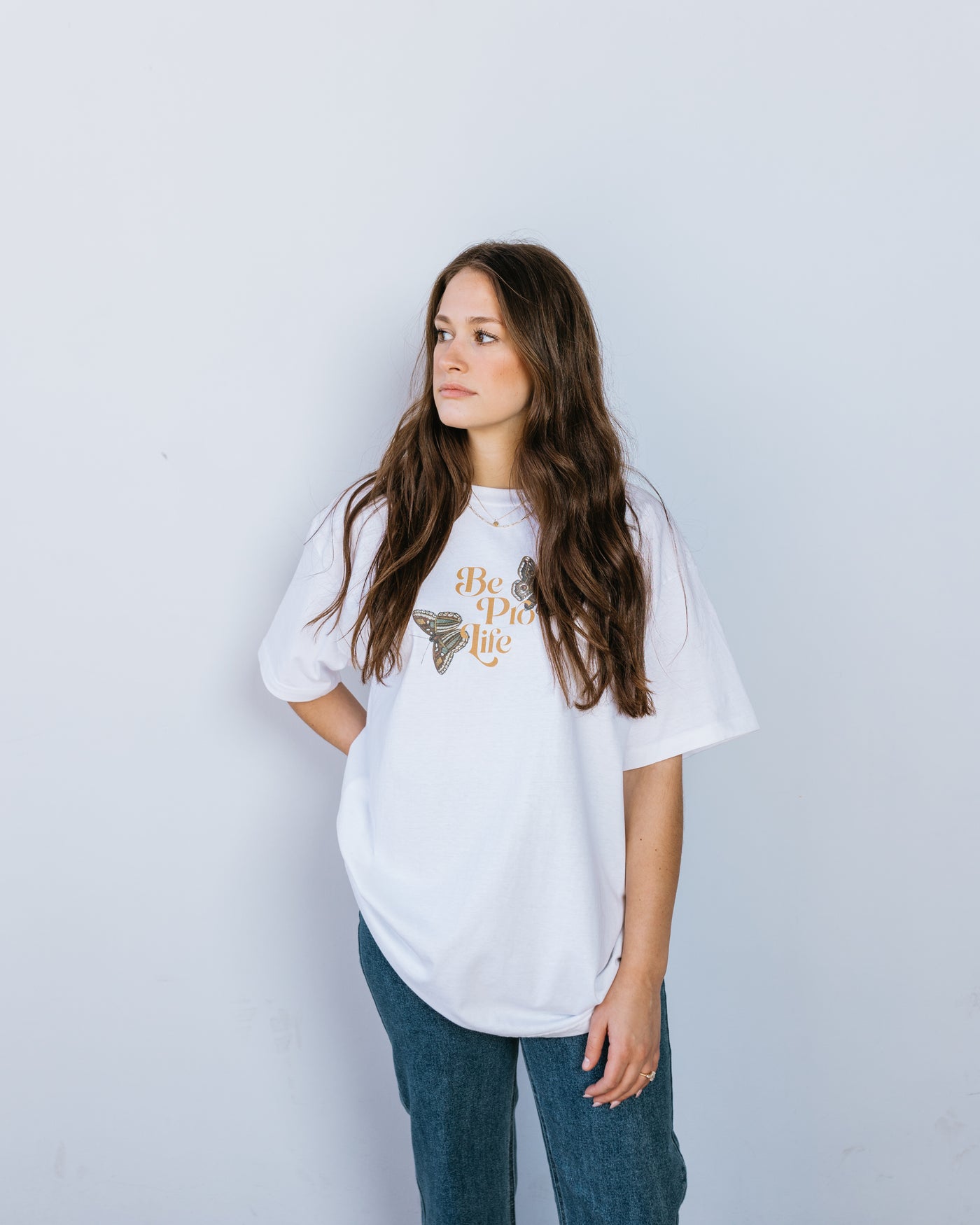 A brunette woman in a white tee shirt with the words "Be Pro Life" in the center surrounded by two large green and brown butterflies.