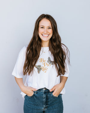 A brunette woman in a white tee shirt with the words "Be Pro Life" in the center surrounded by two large green and brown butterflies.