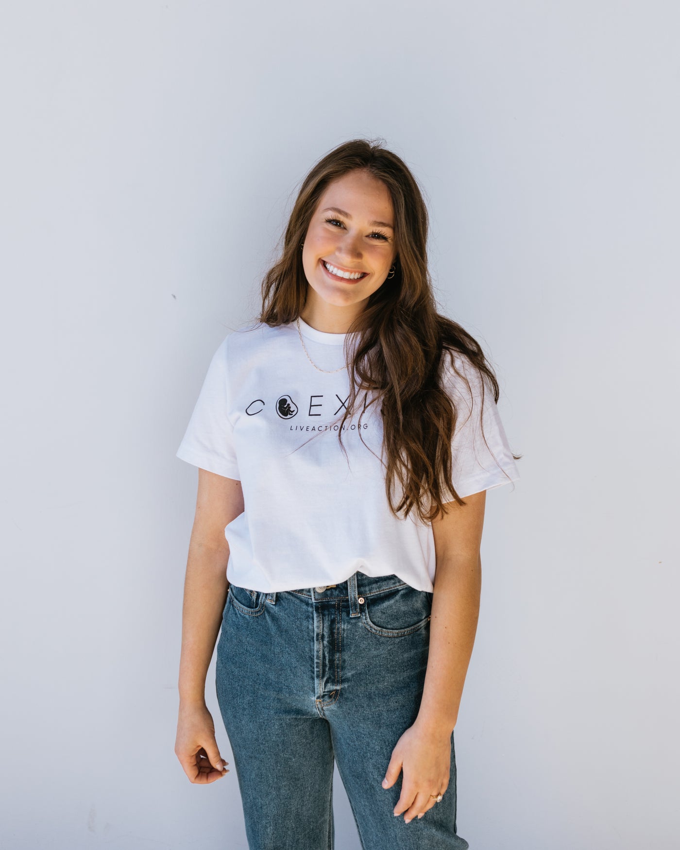 A woman with brown hair in a white tee with the word "COEXIST" written across the chest in fine black font. Inside the misshapen O is a preborn baby. Underneath the main text is Live Action's website written in small font.